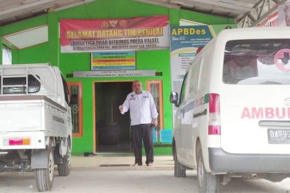 Kepala Desa Penggalaman, Kecamatan Martapura Barat, Kabupaten Banjar, berdiri di depan kantor desa. Sejumlah kendaraan operasional, termasuk ambulans desa, tampak terparkir di halaman kantor. Foto: Rifai/lenterakalimantan.com